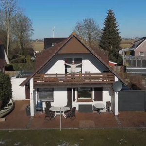 Der Immobilienmakler Sneider steht auf einem Balkon und streckt die Arme begrüßend heraus. Die Immobilie hat ein Braunes Dach und Weiße Mauern